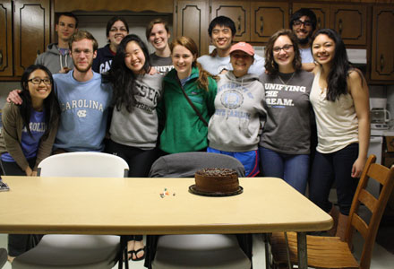 A group of students pose for a photo near a birthday cake. 