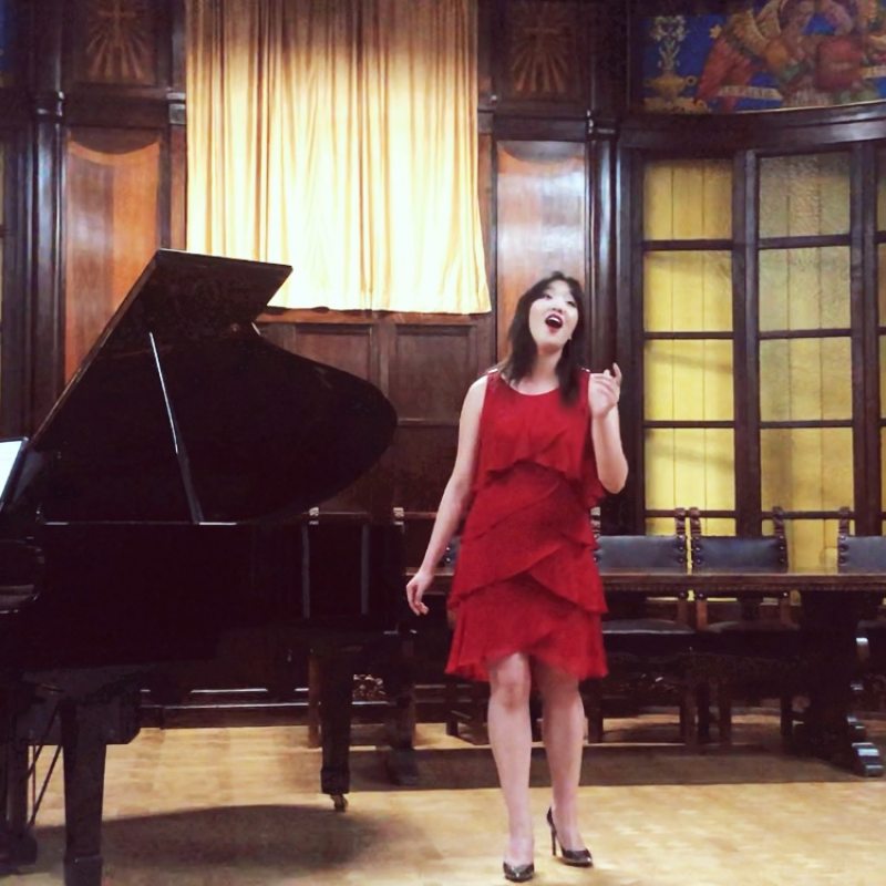 Melody stands beside a grand piano and sings