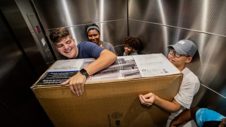 Students ride in an elevator with a futon in a box during move in.