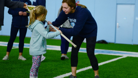 A female lacrosse player hands a lacrosse stick to a young girl.