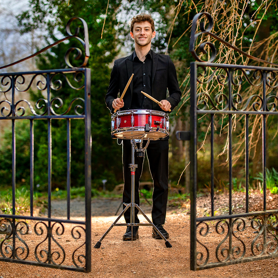 A man plays a snare drum near a fence gate.