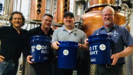 Four men hold blue buckets near distillery equipment