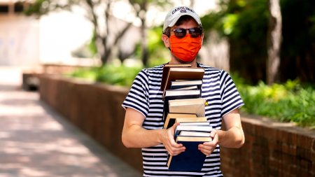 A man wearing an orange mask carries a stack of books on campus.