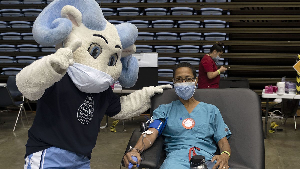 A woman in a mask donated blood next to a mascot of a ram.
