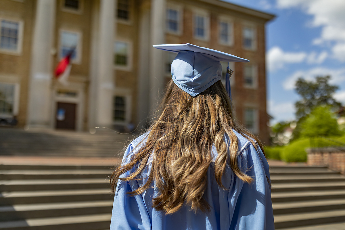 A graduate in a cap and gown stands near South Building.