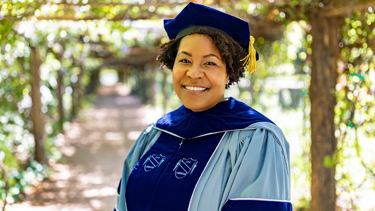 Alexis Dennis in doctoral robes outside in a garden
