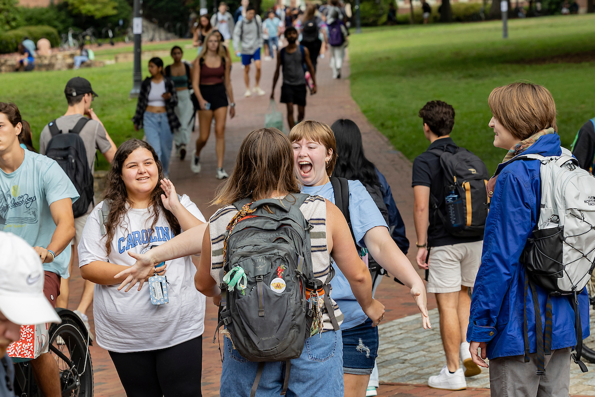 A student smiles as she leans into to hug a friend on campus.