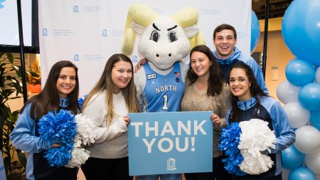 Five students and the Tar Heels mascot Rameses smile at the camera and hold pom poms and a thank you sign