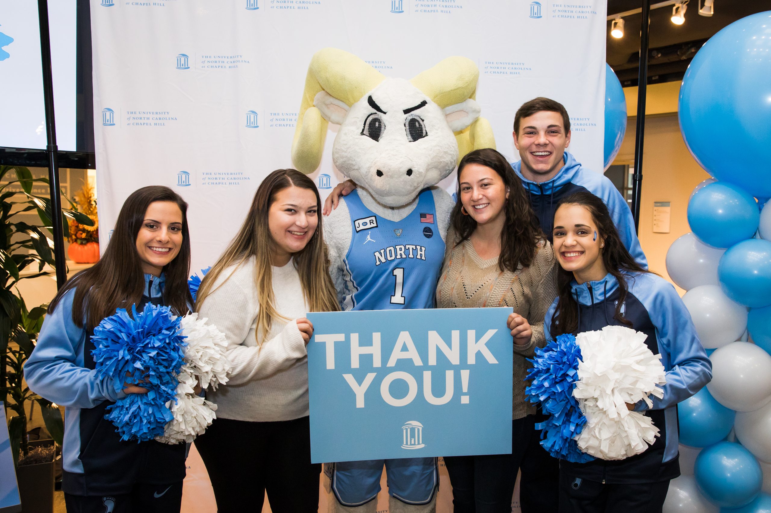 Five students and the Tar Heels mascot Rameses smile at the camera and hold pom poms and a thank you sign