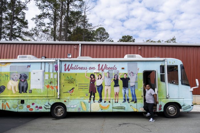 A blue truck with 'Wellness on Wheels" painted on the side along with people painted on the front, parked in front of a brown building with a man standing in the doorway. 