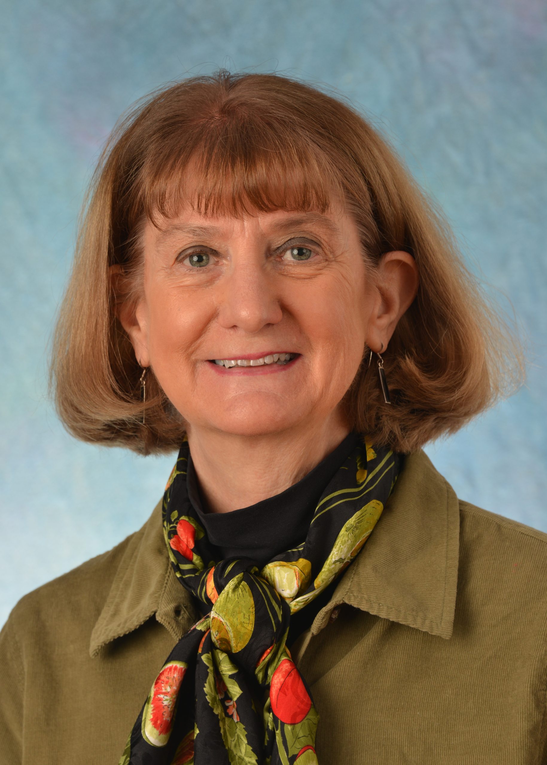 Susan Beck smiling at the camera with a green collared overshirt and a black floral patterned scarf against a blue background.