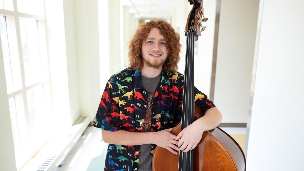 A male student, Sam Bivona, smiling and posing for a photo in a hallway with his arms wrapped around a standup bass instrument.