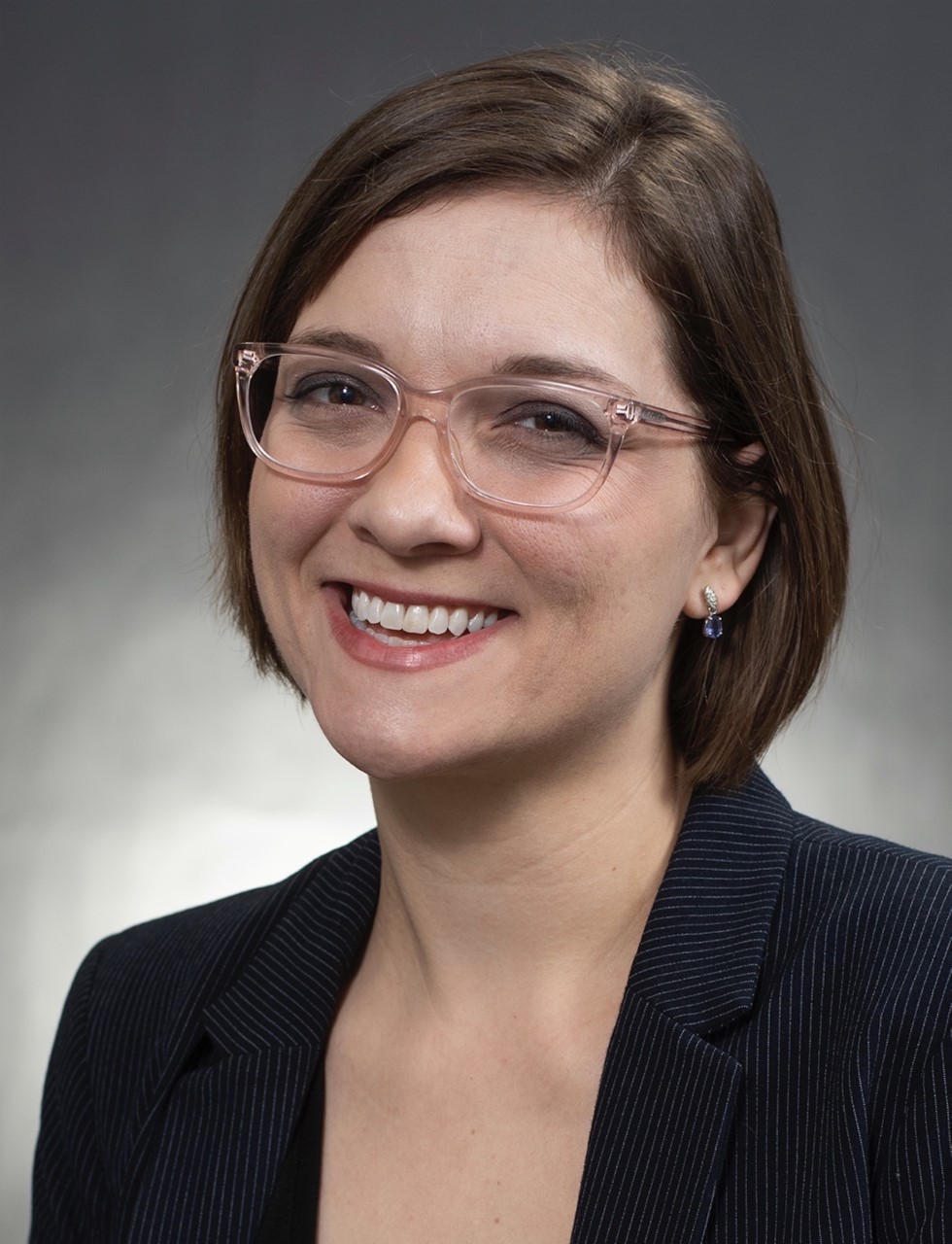 Wendy Clark smiling a the camera in glasses and a black jacket against a background that fades from dark gray on the edges to white in the middle.