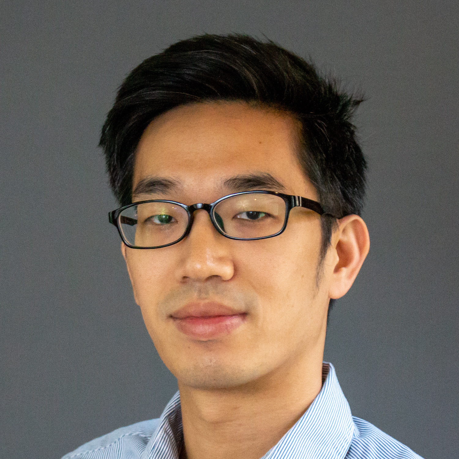 Brian Hsu looking at the camera with black rectangular glasses in a light blue collared shirt against a gray background.