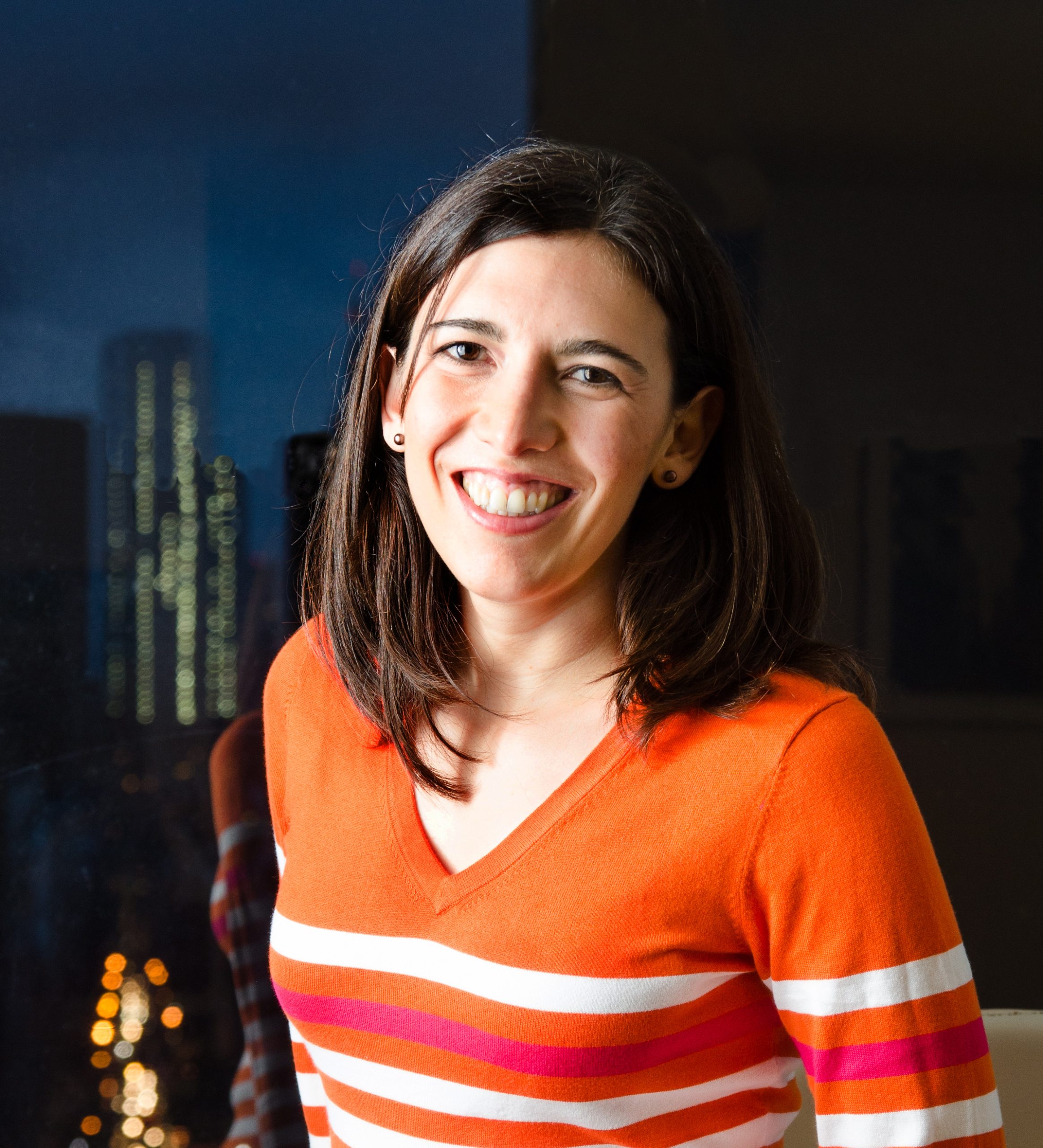 Carla Merino-Rajme smiling at the camera in a bright orange and white striped v-neck shirt with a night-time city skyline in the background.