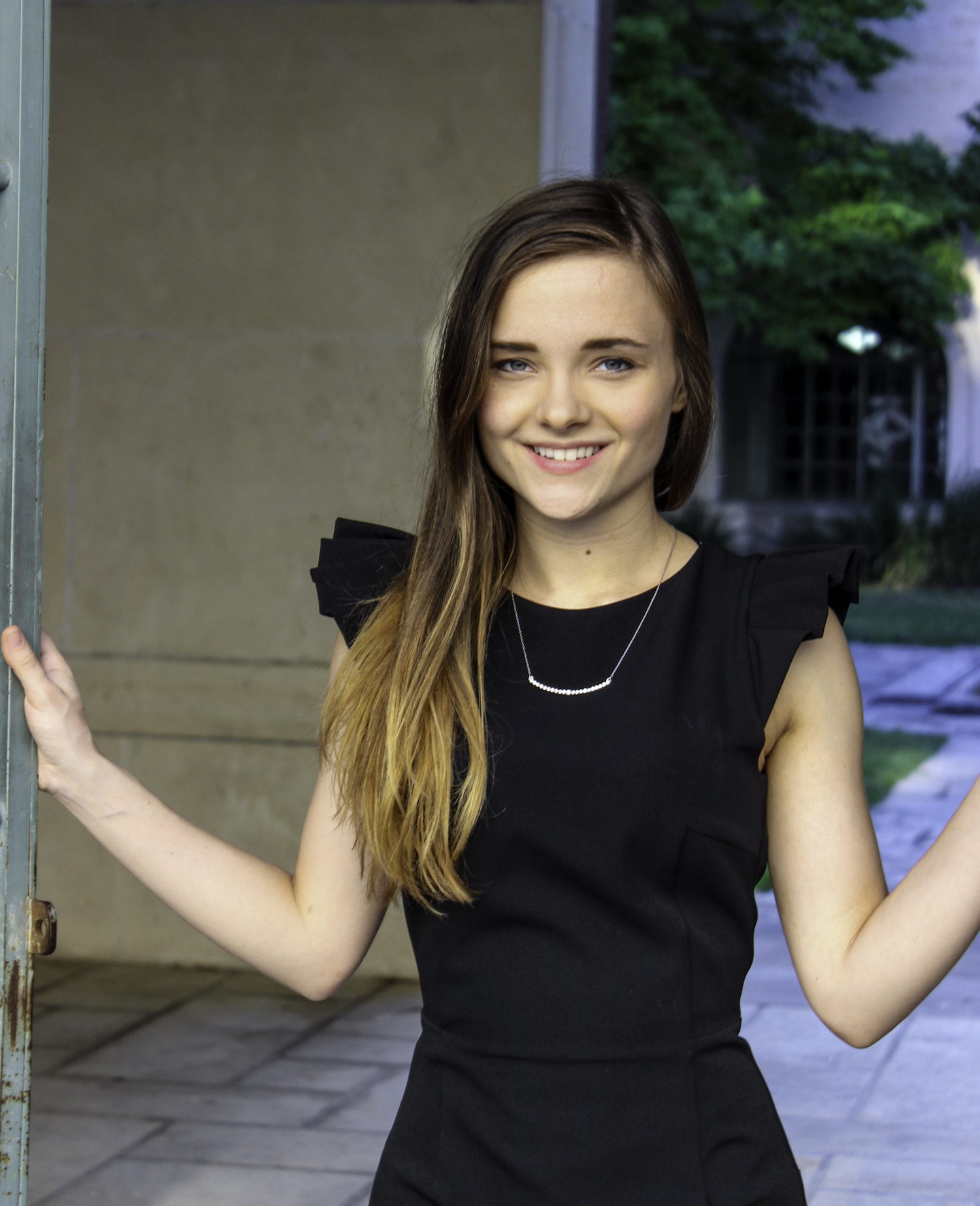 Abigail Newell stands in a black dress with ruffled shoulders, smiling at the camera, with her hands on either side of a doorway in an outdoor space.