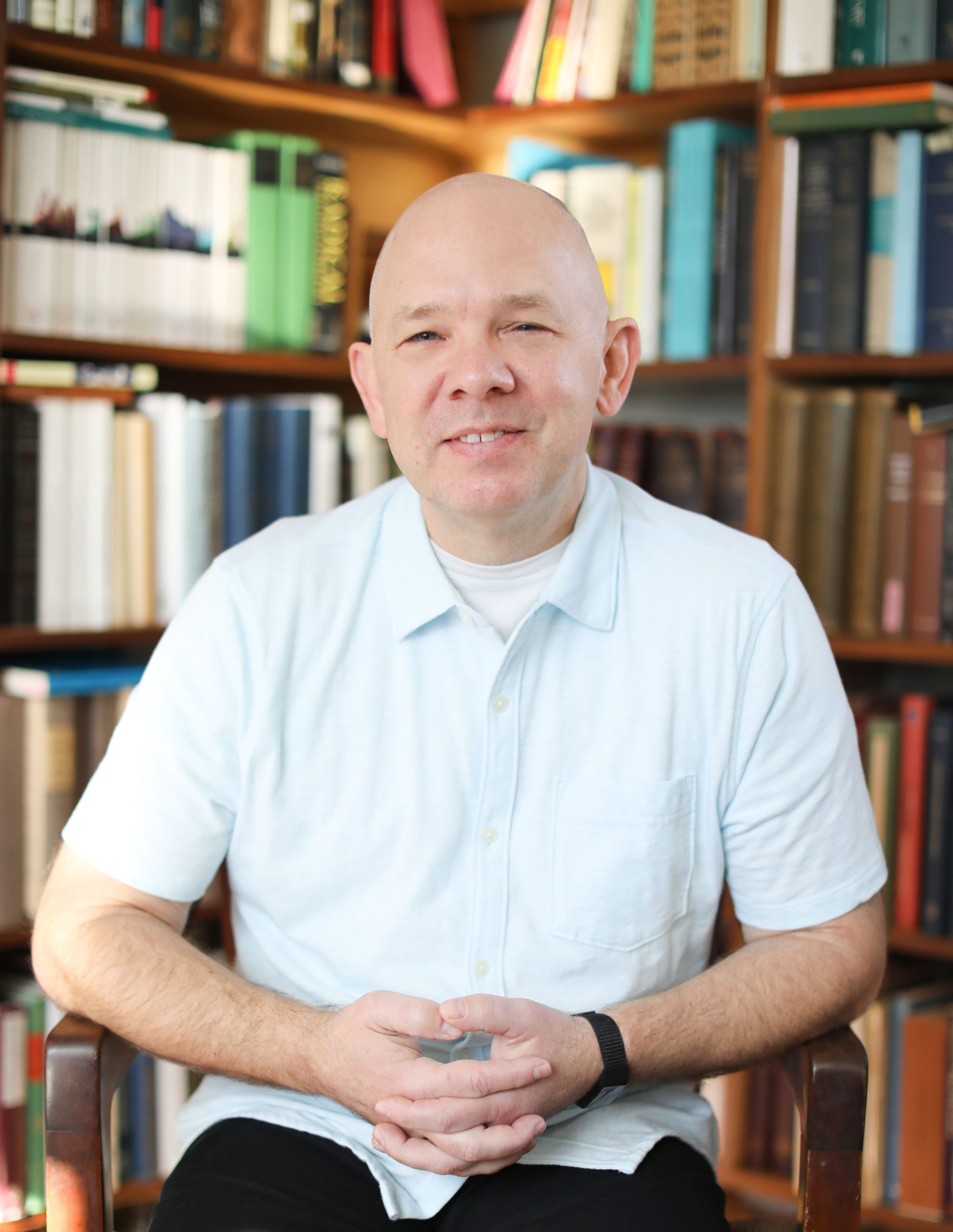 Soren Palmer smiling at the camera in a white polo shirt with bookshelves and books behind him. 