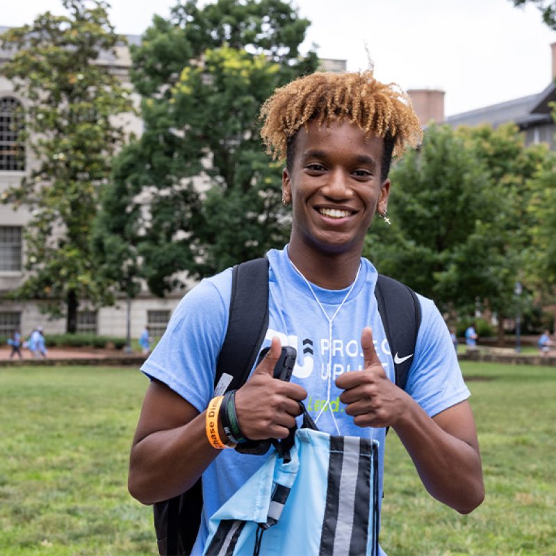 A student smiling and giving a thumbs up while posing for a photo on Polk Place on the campus of UNC-Chapel Hill.
