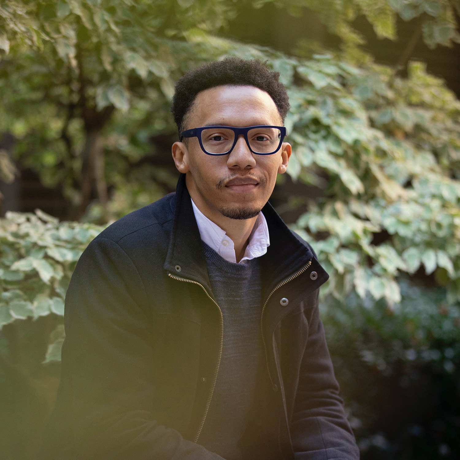 Sidney Wilkerson-Hill looking at the camera in glasses, a dark gray sweater over a light pink collared shirt with a dark overcoat in front of green foliage. A faint green blur is in front of his torso.
