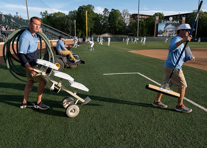 The crew walks out to the infield. Anderson has a hose coiled over his shoulder and others are on a three-wheeled drag and holding a rake. (Jon Gardiner/UNC-Chapel Hill)