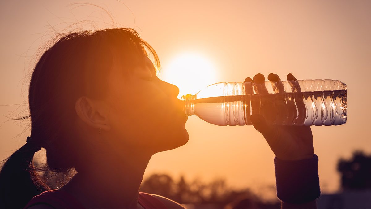 Silhouette of woman drinking from water bottle in front of bright sun and orange sky