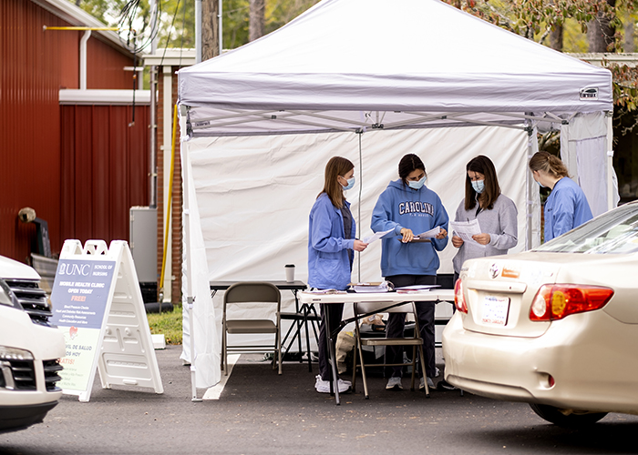 Standing under a tent looking at forms, Students Peyton Gully, Emma Kruger, Moriah Fender and Tara Horoho (left to right) get organized for a UNC School of Nursing mobile nursing unit as cars line up at the CORA Food Pantry.