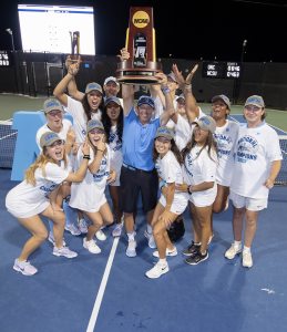 Players from the UNC women's tennis team and coach Brian Kalbas posing for a group picture and lifting a trophy after winning the national championship.