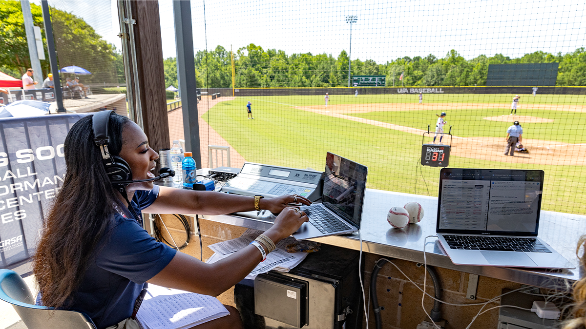 A woman named Jasmine Baker sitting in an open-air press box and providing play-by-play commentary of a baseball game. She is wearing a headset, has a scoresheet sitting in her lap and is also using a laptop to reference notes.