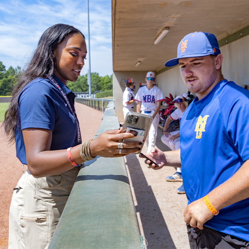 A woman named Jasmine Baker holding a smartphone and showing it to a baseball coach standing in a dugout.