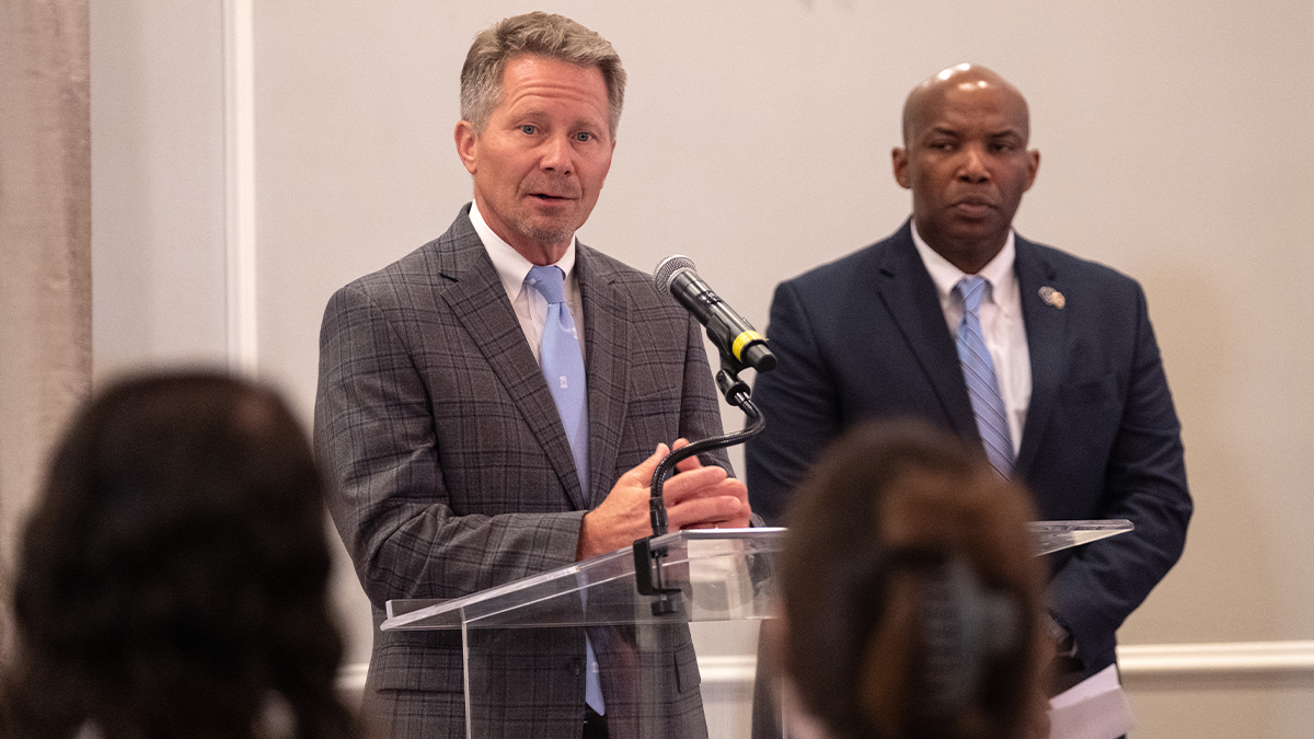 A man, UNC-Chapel Hill Chancellor Kevin M. Guskiewicz, speaking into a microphone at a podium at a news conference. Standing next to him is another man, UNC Police Chief Brian James.