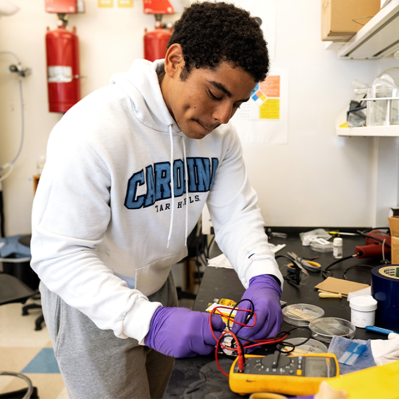A student named Miguel Reyna working in a laboratory, wearing purple gloves and holding red and black wires while conducting research on batteries.