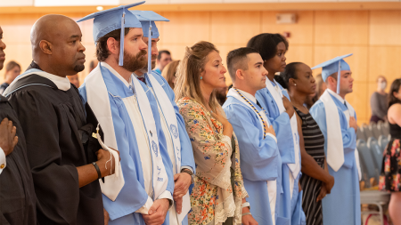 Students in caps and gowns and faculty members standing and placing their hands over their hearts.