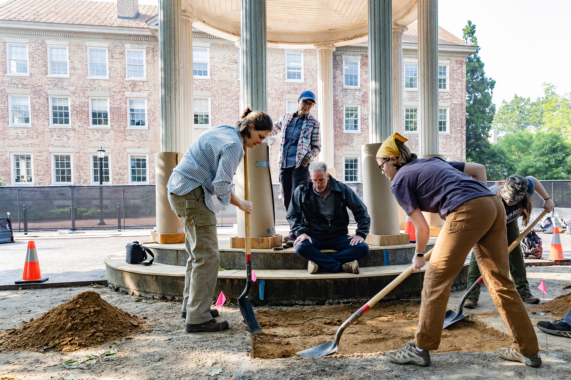 Two students using shovels to dig into the ground next to a well. Two onlookers are in the background.