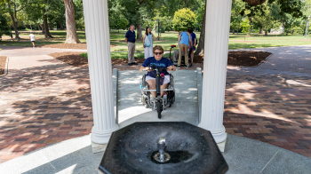 A person using a wheelchair on a sloped pathway approaches the fountain of the Old Well on the campus of UNC-Chapel Hill.
