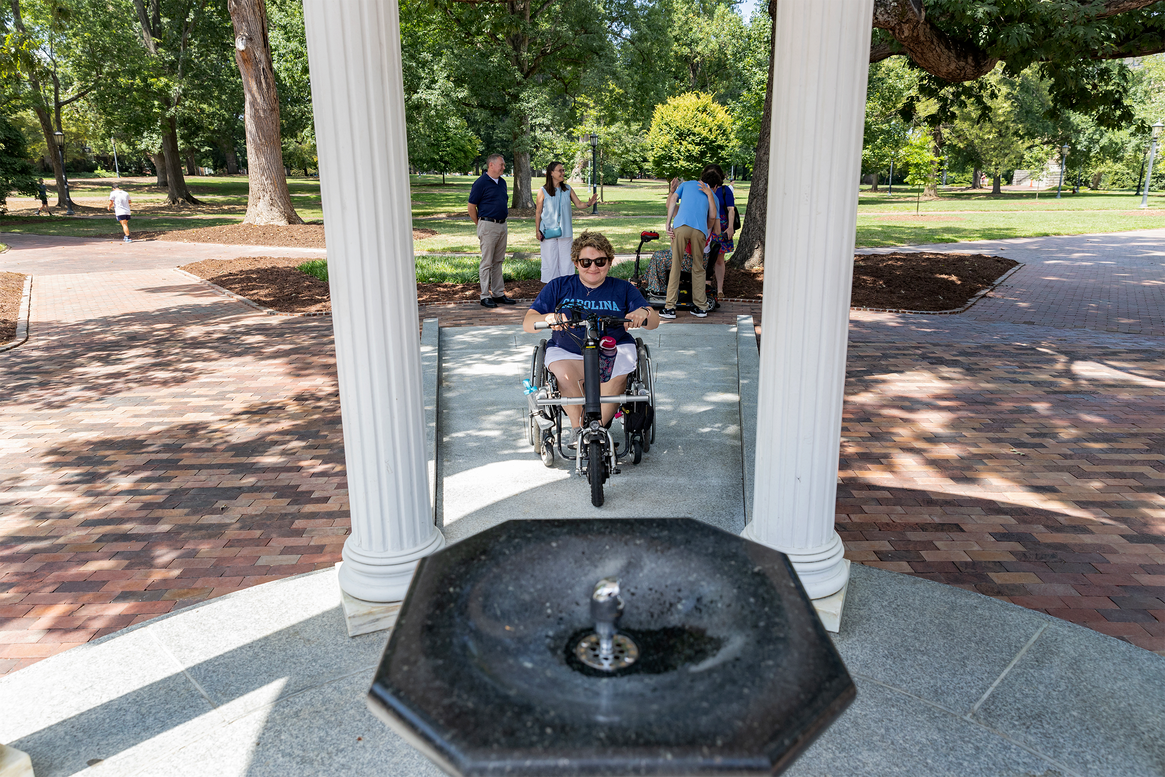 A person using a wheelchair on a sloped pathway approaches the fountain of the Old Well on the campus of UNC-Chapel Hill.