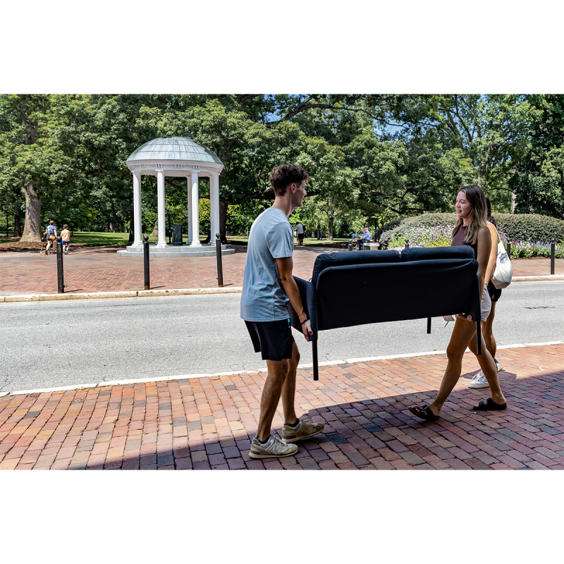 Two students carrying a couch on a sidewalk on the campus of UNC-Chapel Hill across the street from the Old Well.