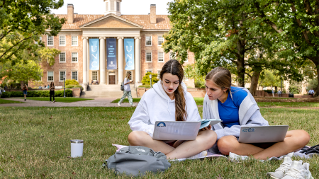 Two female students sitting on a lawn and looking at a laptop on the campus of UNC-Chapel Hill.
