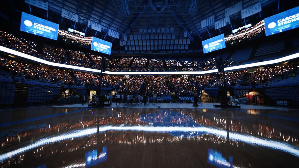 Wide-angle shot of a large crowd of people holding up candles in a dimly lit arena during a vigil. Four large video boards displaying graphics reading 
