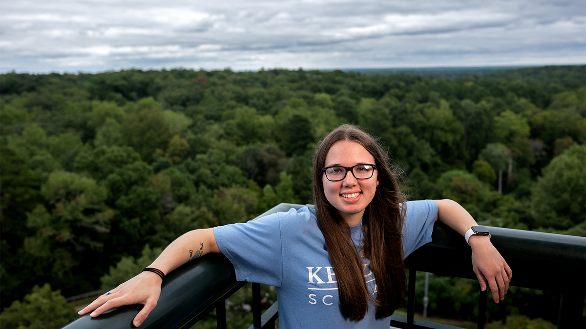 A student, Emily Shipway, posing for a photo on the balcony of a dormitory with a gray sky and an aerial view of green trees in the horizon.