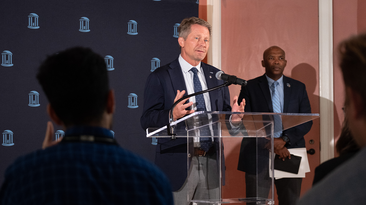 A man, UNC-Chapel Hill Chancellor Kevin M. Guskiewicz, speaking into a microphone at a dais in a news conference. Standing in the background is another man, UNC Police Chief Brian James.