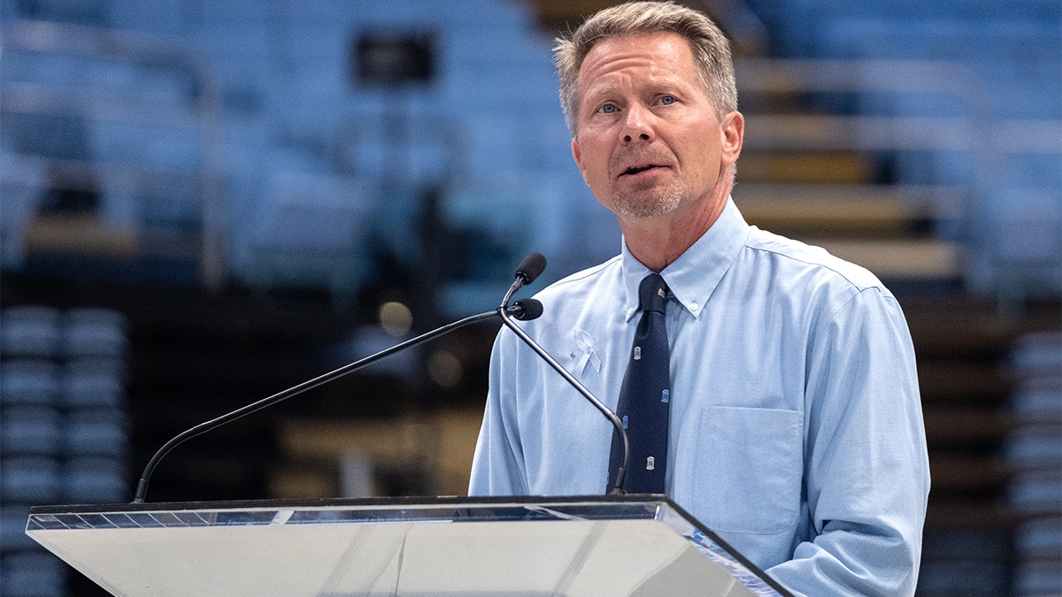 A man, UNC-Chapel Hill Chancellor Kevin M. Guskiewicz, speaking into a microphone at a podium in an arena while addressing a crowd at a vigil.