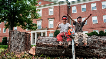 Two male workers smiling and posing for a photo sitting atop a chopped down tree in front a brick building.