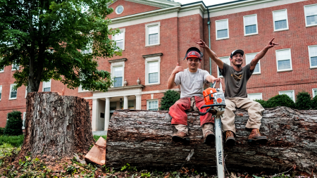Two male workers smiling and posing for a photo sitting atop a chopped down tree in front a brick building.
