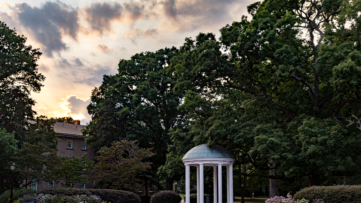 View an the Old Well on the campus of UNC-Chapel Hill at dusk with trees, flowers and bushes surrounding the structure.