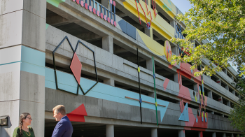 A woman and a man talking in front of a parking deck that has an art installation on its exterior.