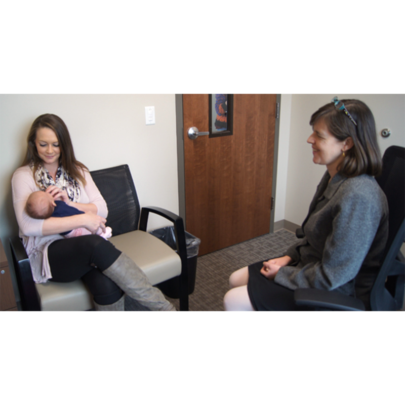 Woman smiling while holding a baby in a chair while talking with another woman inside of an office.