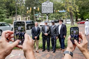 A group of four men standing and posing for a photo in front of a newly unveiled historical marker on the side of Franklin Street. In the foreground, you can see hands holding iPhones to take pictures.
