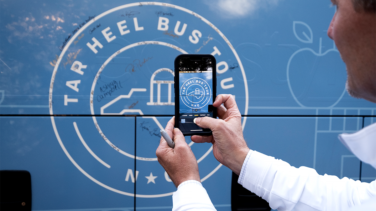 A man taking a photo on an iPhone of the side of a bus that features the logo of the Tar Heel Bus Tour on its side.