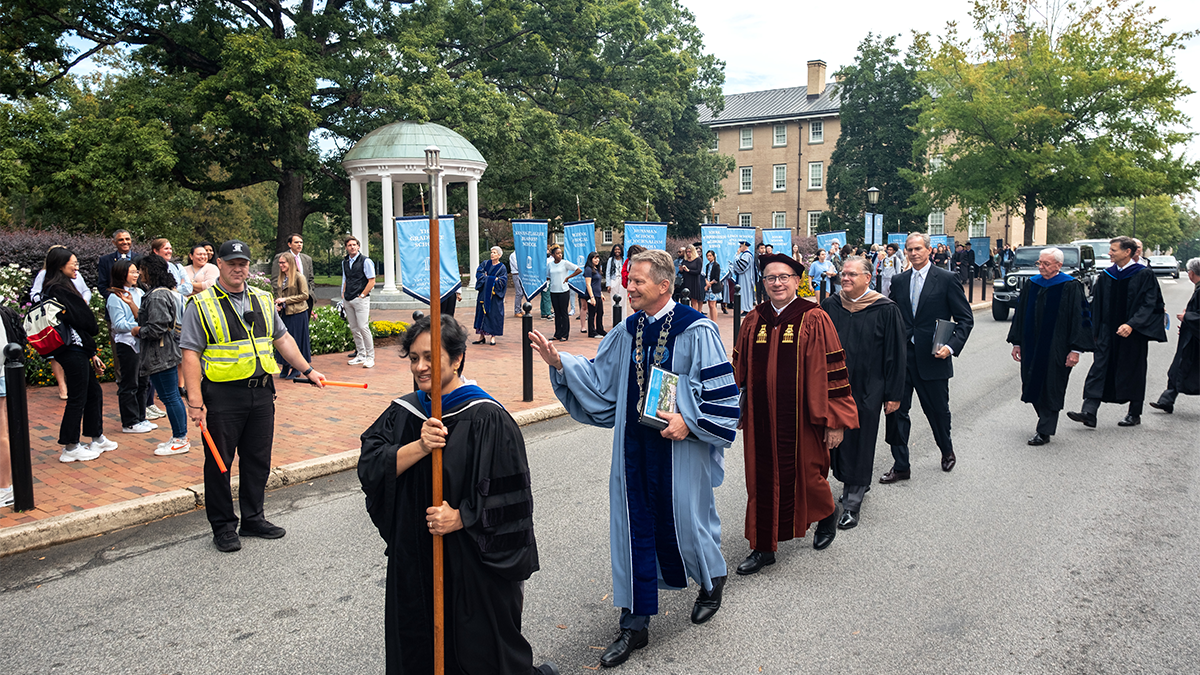 Men and women wearing ceremonial robes walk across a street near the UNC's Old Well.