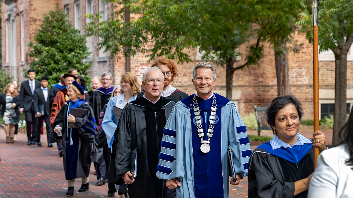 University leaders in regalia walking on a brick pathway on the campus of UNC-Chapel Hill as part of a past University Day celebration. Smiling at the camera is Chancellor Kevin M. Guskiewicz.